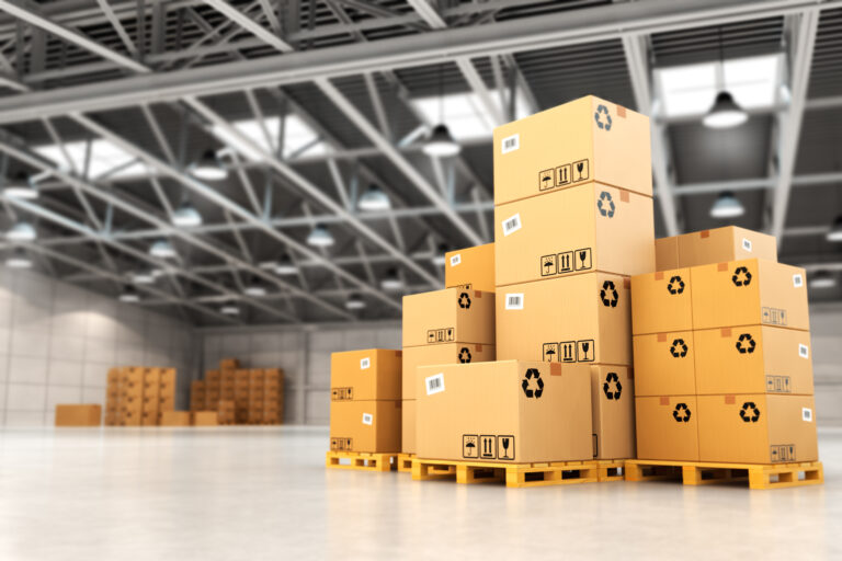Cardboard boxes stacked on a wooden pallet inside a spacious warehouse with high shelves and industrial lighting.