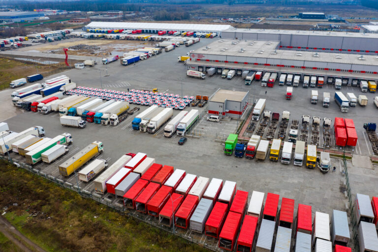 Aerial drone photo of a large logistics yard with hundreds of trucks and trailers parked in organized rows, alongside a warehouse with multiple loading docks